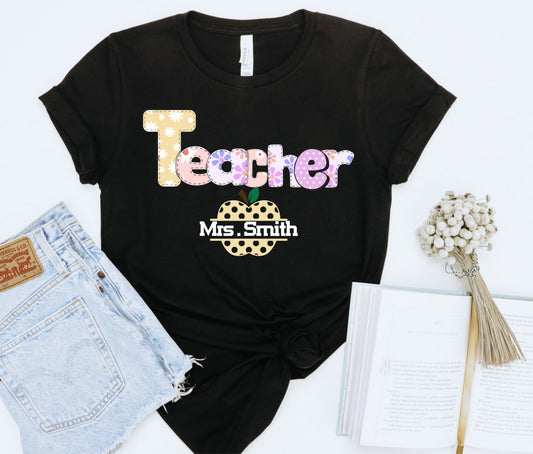 Black t-shirt with 'Teacher' and apple design, paired with denim shorts and a book on a white background